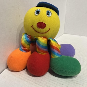 May include: A plush octopus toy with a bright yellow round head, a small red nose, and blue eyes. It wears a blue cap and features eight tentacles, each with a vibrant rainbow-striped pattern and solid-colored ends in orange, red, and green.