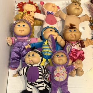 May include: A collection of eight Cabbage Patch Kids dolls with various outfits, including a lion costume, a pink pig suit, a purple glittery jumpsuit, a blue patterned outfit, a zebra costume, and a purple bunny suit, all with distinct facial features.