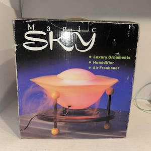May include: A box for a "Magic Sky" humidifier and air freshener. The product is a light orange, saucer-shaped device with a white top, supported by black metal legs and a circular base. The box has the words "Magic Sky" in white.