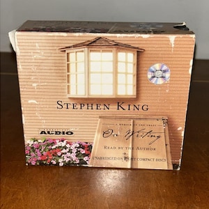 Puede incluir: Caja de audiolibro de Stephen King titulada "On Writing: A Memoir of the Craft". La caja presenta una ilustración de una casa con mirador y un jardín de flores. El set incluye discos compactos.