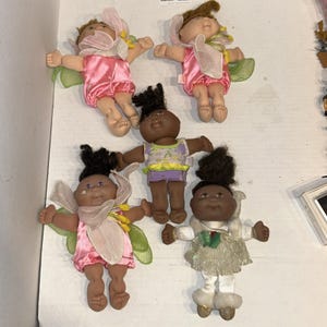 Puede incluir: Una colección de cinco muñecas pequeñas con cuerpos blandos y cabezas de vinilo. Dos muñecas llevan trajes rosas con alas transparentes, una tiene una parte superior morada y amarilla con pantalones cortos, una tiene ropa rosa con alas verdes y una vestido.