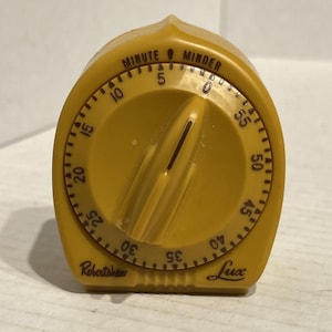 May include: A vintage, mustard-yellow kitchen timer with a rounded, arched design. The timer features a large dial with minute markings and a central knob. The words "MINUTE MINDER", "Robertshaw", and "Lux" are visible.