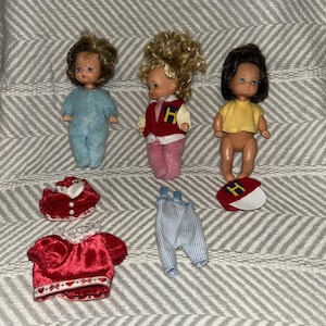 May include: Three vintage doll figures with various outfits. One doll wears a blue jumpsuit, another a red jacket with a white collar and pink trousers, and the third a yellow top. Additional clothing includes a red hat and striped dungarees.