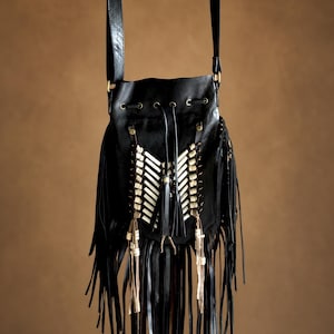May include: A black drawstring shoulder bag featuring long fringe detailing along the bottom and sides. The front is decorated with a central panel of white bone-like tubes arranged in a chevron pattern, accented by brass-toned metal hardware.