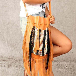 May include: A tan drawstring bucket bag featuring long fringe trim along the sides and bottom. The front panel is decorated with a vertical arrangement of white cylindrical beads held in place by black cord lacing and a matching tassel.
