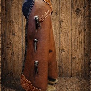 May include: Brown leather chaps with decorative silver conchos and black fringe. The chaps feature detailed stitching and tooling along the edges and are designed for riding. The background is a rustic wooden wall.