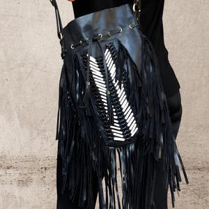 May include: A black bucket bag featuring long fringe detailing, a drawstring closure with brass-toned metal eyelets, and a central decorative panel adorned with white cylindrical beads and black woven cord accents, suspended by a shoulder strap.