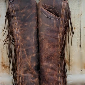 May include: Brown leather chaps with decorative floral tooling on the yoke. The chaps have fringe along the sides and decorative stitching. A pocket is visible on the right leg. The chaps are hanging against a wooden background.