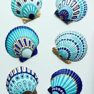 Może przedstawiać: Sześć ręcznie malowanych muszli przegrzebków w odcieniach niebieskiego, turkusowego i złotego. Każda muszla ma unikalny wzór z geometrycznymi wzorami. Muszle są ułożone na białym tle, prezentując ich skomplikowane detale.