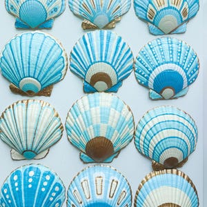 Pode incluir: Uma variedade de conchas pintadas em tons de azul, branco e dourado. As conchas apresentam vários padrões, incluindo listras e pontos, com detalhes dourados. As conchas estão dispostas sobre uma superfície branca.
