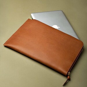 以下が含まれることがあります： 茶色の革製ラップトップスリーブ、ジッパー開閉式。 長方形のスリーブは滑らかな質感です。 Appleのロゴが入ったシルバーのラップトップがスリーブの後ろに部分的に見えています。