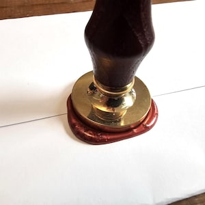 Peut inclure: Gros plan d'un sceau de cire. Le tampon est composé d'un manche en bois brun foncé, d'une base en métal doré et d'un sceau de cire rouge. Le tampon est pressé sur une surface blanche.