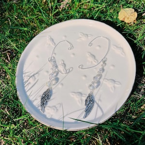May include: A pair of silver ear cuffs with a wing and rose charm. The ear cuffs are adorned with clear beads and are displayed on a white plate with a floral design, set against a backdrop of green grass.