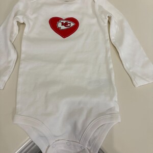 May include: White baby bodysuit with long sleeves. Features a red heart with the Kansas City Chiefs logo in the center. The bodysuit is made of soft material and has snap closures at the bottom. The brand name "Carter's" is printed on the top.