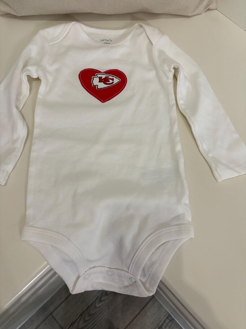 May include: White baby bodysuit with long sleeves. Features a red heart with the Kansas City Chiefs logo in the center. The bodysuit is made of soft material and has snap closures at the bottom. The brand name "Carter's" is printed on the top.