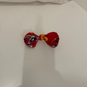 May include: A red bow tie with a repeating pattern of a race car in black and white. The center knot is yellow. The bow tie is on a white surface.
