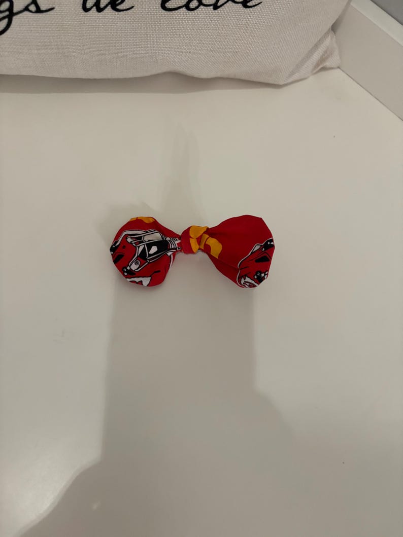 May include: A red bow tie with a repeating pattern of a race car in black and white. The center knot is yellow. The bow tie is on a white surface.