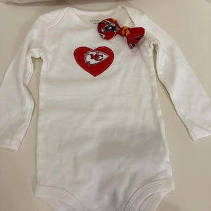 May include: White long-sleeved baby bodysuit with a red heart-shaped patch featuring a yellow and white logo. A matching red bow is attached. The bodysuit is a Carter's brand.