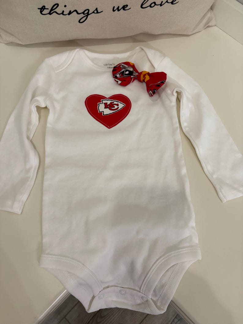May include: White long-sleeved baby bodysuit with a red heart-shaped patch featuring a yellow and white logo. A matching red bow is attached. The bodysuit is a Carter's brand.