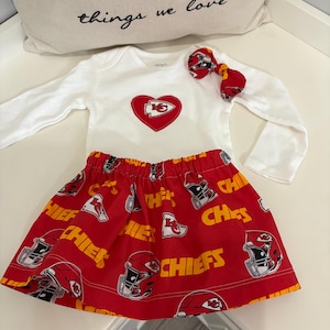 May include: A white long-sleeved baby bodysuit with a red heart-shaped patch featuring a team logo. The red skirt has a repeating pattern of helmets and the word "CHIEFS". A matching bow completes the outfit.