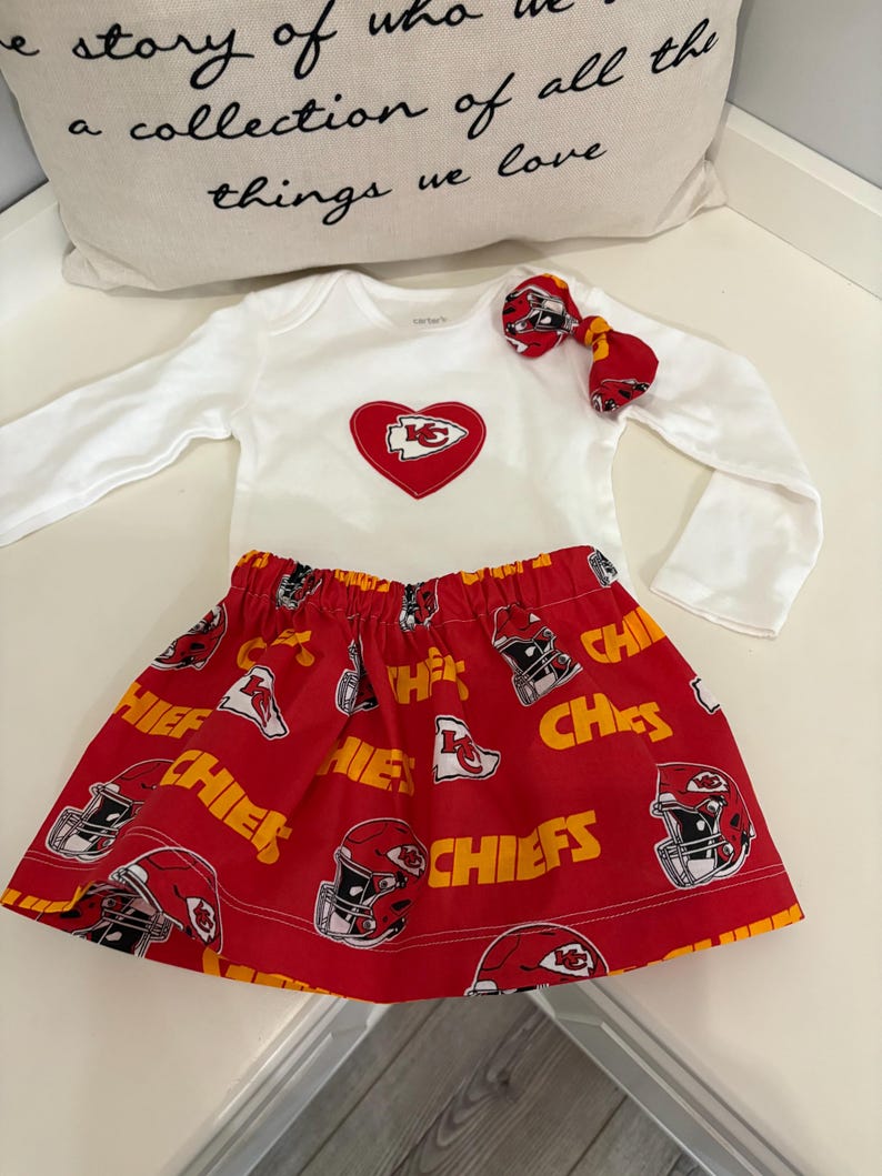 May include: A white long-sleeved baby bodysuit with a red heart-shaped patch featuring a team logo. The red skirt has a repeating pattern of helmets and the word "CHIEFS". A matching bow completes the outfit.