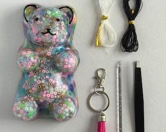 May include: A clear resin bear figurine filled with multicolored beads, accompanied by a white cord, a black cord, a silver keychain with a pink tassel, a clear plastic tool, and a pair of black tweezers for crafting and assembly.