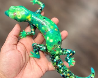 May include: A translucent green lizard figurine crafted from resin, featuring an embedded pattern of multicolored beads and small circular rings throughout its body, including its head, four legs, and a long, curved tail with a smooth, glossy finish.