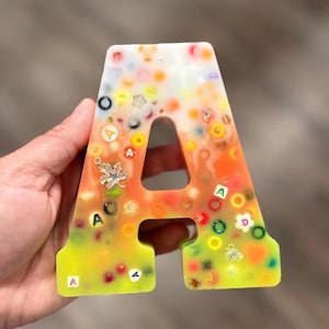 May include: A letter A shaped resin ornament featuring a gradient color transition from white at the top to orange in the middle and yellow at the bottom. The piece is embedded with various colorful beads, small letter cubes, and metallic charms.