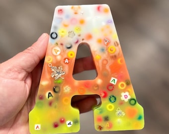 May include: A letter A shaped resin ornament featuring a gradient color transition from white at the top to orange in the middle and yellow at the bottom. The piece is embedded with various colorful beads, small letter cubes, and metallic charms.