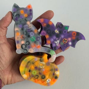 May include: A collection of Halloween-themed resin figurines. Includes a gray cat with a skeleton design, a purple bat, and an orange pumpkin. Each piece has colorful accents and small metal charms. The cat measures approximately 4 inches in height.