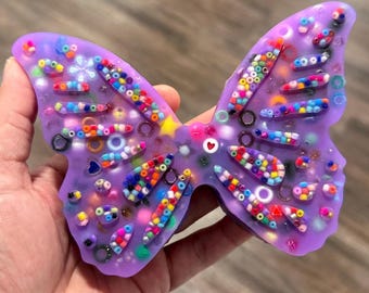 May include: A butterfly-shaped object made of translucent purple resin, embedded with a variety of colorful multi-shaped beads, small rings, and a single white heart-shaped bead with a red center, creating a textured and vibrant surface.