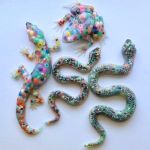 May include: A collection of four animal figurines made of translucent resin embedded with colorful beads. The set includes a lizard, a frog, and two snakes, each featuring a unique arrangement of multi-colored plastic beads throughout their bodies.