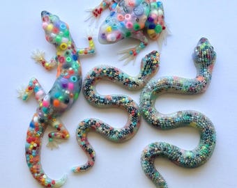 May include: A collection of three translucent animal figurines including a lizard, a frog, and a snake. The lizard and frog are embedded with large, multi-colored plastic beads, while the snake is filled with tiny, dark, multi-colored beads.