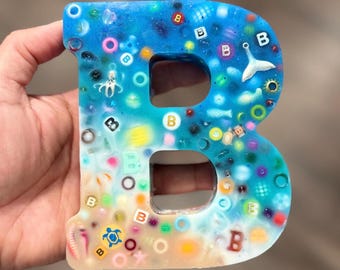May include: A letter B shaped resin ornament featuring a blue to white gradient. The surface is embedded with various colorful beads, small letter cubes, a white whale tail charm, and a yellow turtle charm, creating a textured, multi-colored appearance.