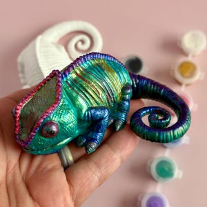 May include: A vibrant, hand-painted chameleon figurine in shades of teal, blue, and purple. The figurine has a curled tail and is held in a hand. Paint pots are visible in the background. The chameleon is a unique piece of art.