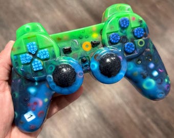 May include: A video game controller featuring a translucent resin shell with a gradient of bright green at the top transitioning to deep blue at the bottom. The device includes blue directional buttons, two black textured joysticks, and a white square button with the number 1 printed on it.