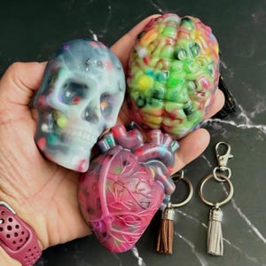 May include: A collection of resin keychains in the shape of a skull, brain, and heart. The skull is light blue with colorful accents. The brain is multi-colored, and the heart is red and pink. Each keychain has a tassel.