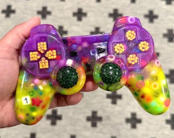Op de afbeelding: Een aangepaste videogamecontroller met een doorschijnende paarse en gele harsbehuizing, ingelegd met kleurrijke kralen, stervormige accenten en kleine cirkelvormige ringen. De controller bevat twee donkergroene, getextureerde joysticks en een wit label met het cijfer 1.