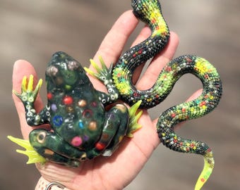 Puede incluir: Una figura de rana verde oscuro con manchas circulares multicolores y patas palmeadas de color amarillo brillante, junto a una figura de serpiente larga y sinuosa con una superficie texturizada de cuentas negras, verdes y amarillas.