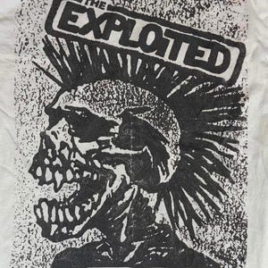Pode incluir: Camiseta branca com um gráfico preto e branco de uma caveira com moicano e o texto "THE EXPLOITED" em uma caixa retangular. O design tem uma estética punk rock desgastada.