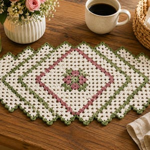 May include: A rectangular crochet doily featuring a diamond-shaped center with pink and green floral motifs, surrounded by layers of white and green yarn in a classic granny square pattern with a scalloped, decorative edge throughout the piece.