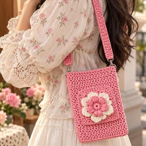 May include: A rectangular crochet crossbody bag in a vibrant pink yarn, featuring a prominent three-dimensional flower motif on the front flap with cream-colored outer petals and a pink center, attached to a matching pink crocheted shoulder strap.