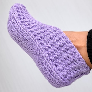 May include: A light purple crochet slipper with a textured, ribbed stitch pattern. The slipper features a rounded toe and a fitted ankle opening, crafted from yarn with a consistent lavender hue throughout the entire piece.