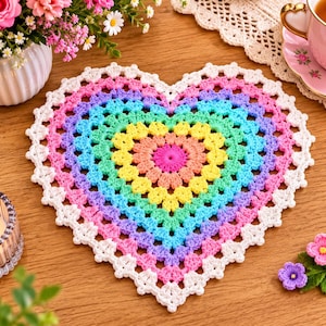 Op de afbeelding: Een hartvormig gehaakt kleedje met concentrische ringen in levendige regenboogkleuren zoals roze, oranje, geel, groen, blauw en paars, afgewerkt met een delicate witte geschulpte kanten rand rondom de gehele buitenste rand.