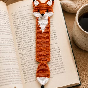May include: Crocheted fox bookmark in orange, white, and black, placed in an open book. The bookmark features a fox head, body, and tail with a black tassel. The book's pages are aged.