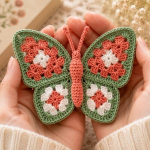 Könnte beinhalten: Ein handgefertigter Häkel-Schmetterling in Grün-, Korallen- und Weißtönen. Die Flügel des Schmetterlings weisen ein Blumenmuster auf, und der Körper und die Antennen sind in passender Korallenfarbe gehalten. Der Schmetterling wird in offenen Händen gehalten.