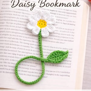 May include: Crochet daisy bookmark on a book page. The bookmark features a white daisy with a yellow center and a green stem and leaf. Text on the page reads "Daisy Bookmark" and "Beginner Friendly • PDF Download."