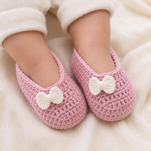 May include: A pair of light pink crocheted baby booties featuring a textured yarn construction and a small white crocheted bow accent on the front of each shoe. The booties are designed with a rounded toe and a simple slip-on style.