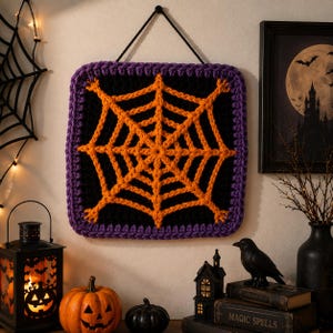 May include: A square crochet wall hanging featuring a vibrant orange spiderweb design centered on a black background, framed by a thick purple border. The piece is suspended by a black cord attached to the top corners for easy wall display.