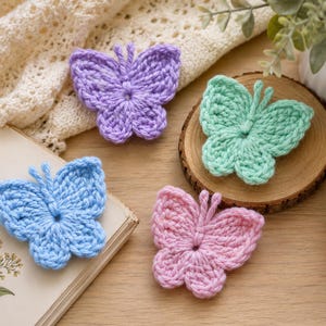 Pode incluir: Quatro borboletas de crochê em cores pastel: uma roxa clara, uma verde menta, uma azul clara e uma rosa clara. Cada borboleta apresenta duas asas superiores, duas asas inferiores e duas pequenas antenas feitas de fio.